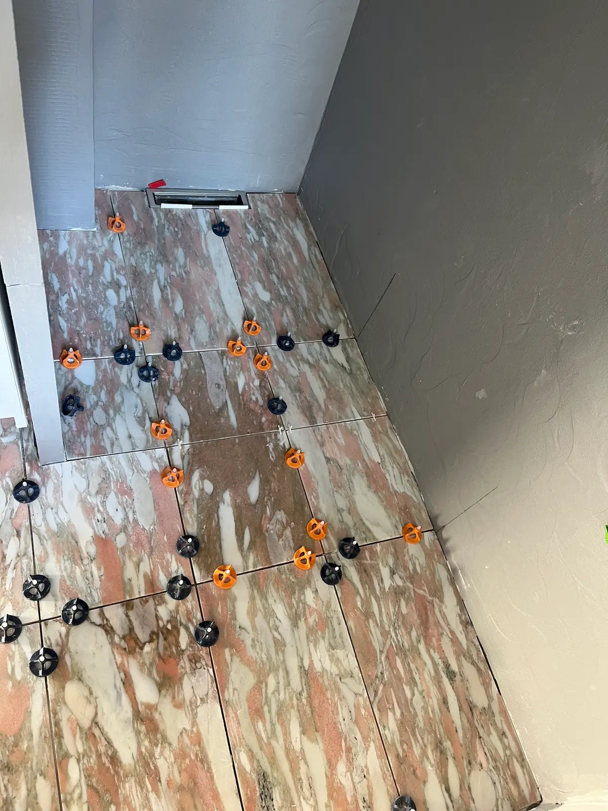 Store rosa marmorfliser lægges på gulvet med orange og sorte nivelleringsklips omkring afløbsrenden