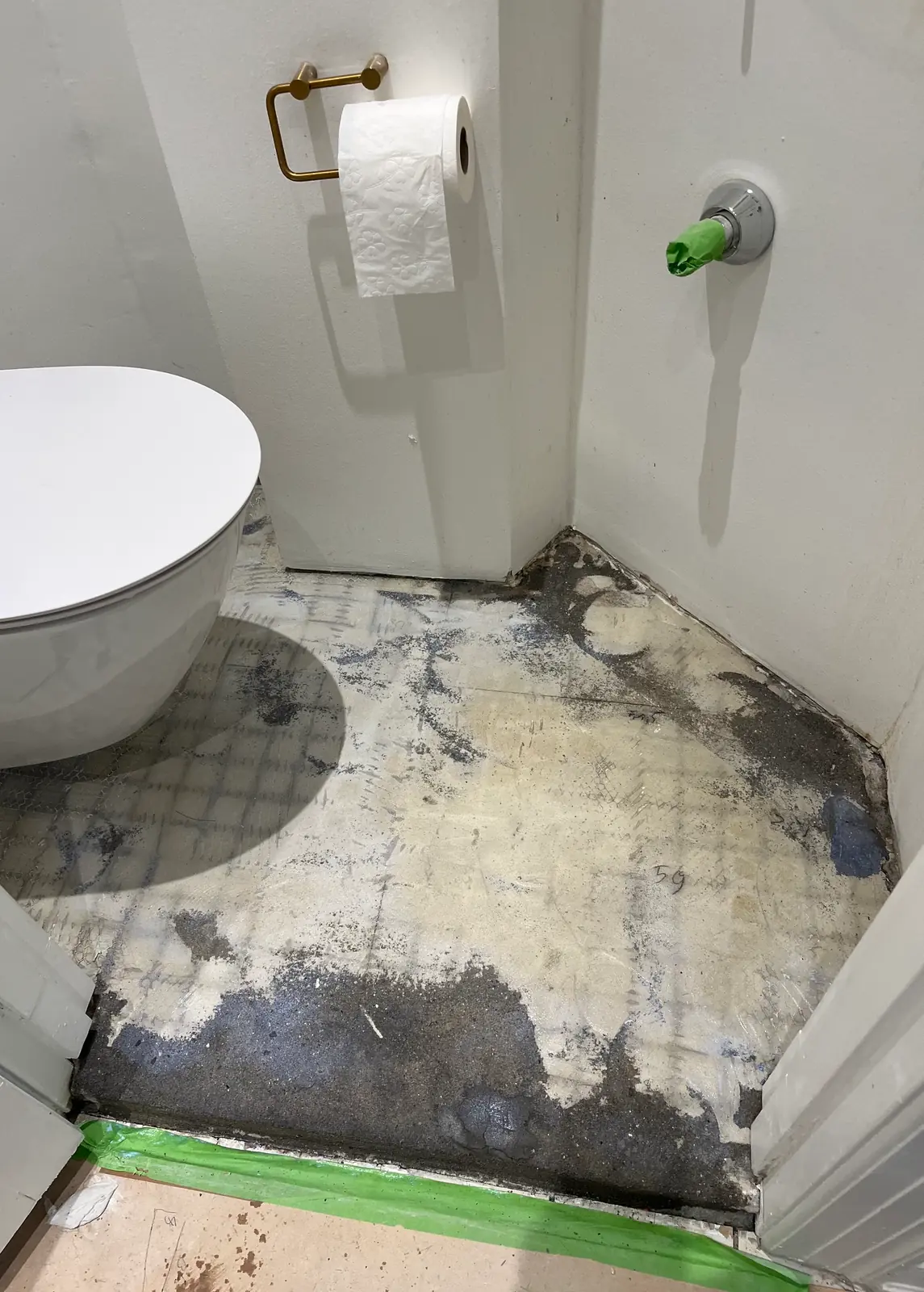 Toiletkrog med bart, plettet undergulv af små gamle fliser, væghængt toilet til venstre, messingholder med papir på væggen og grøn tapelinje langs soklen