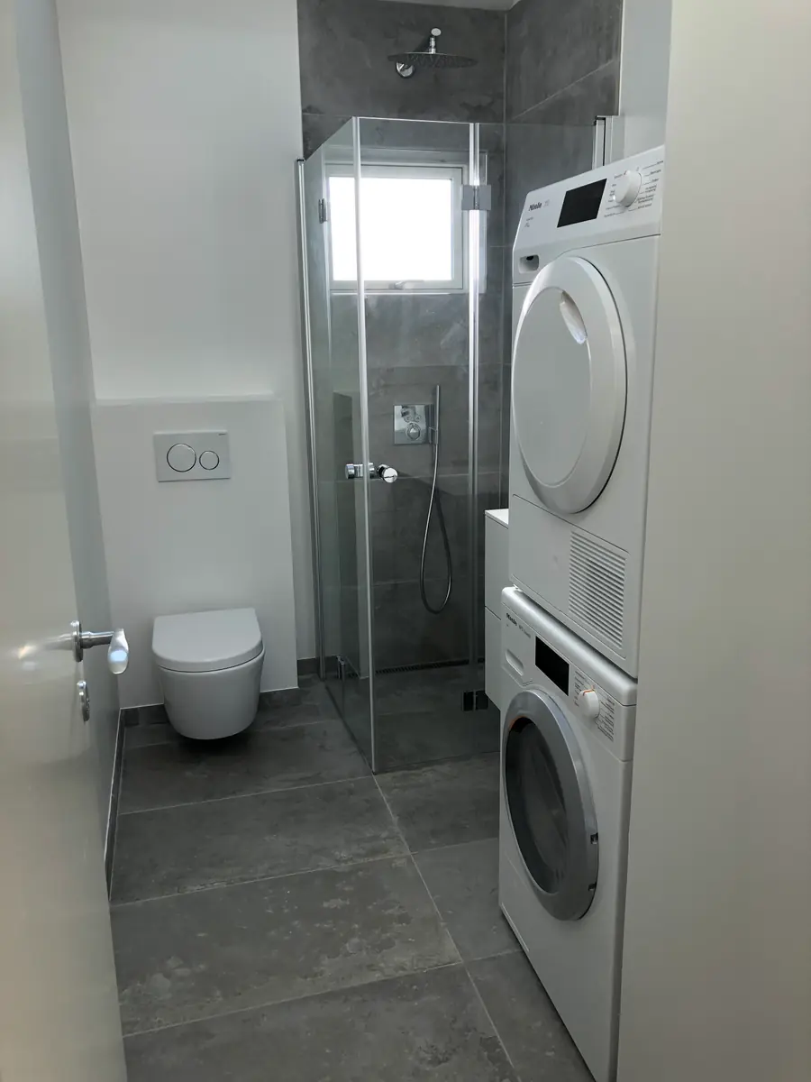 Smalt badeværelse med gråt flisegulv, grå storformatfliser i brusekabinen bag glasdør, væghængt toilet og vaskesøjle med vaskemaskine og tørretumbler