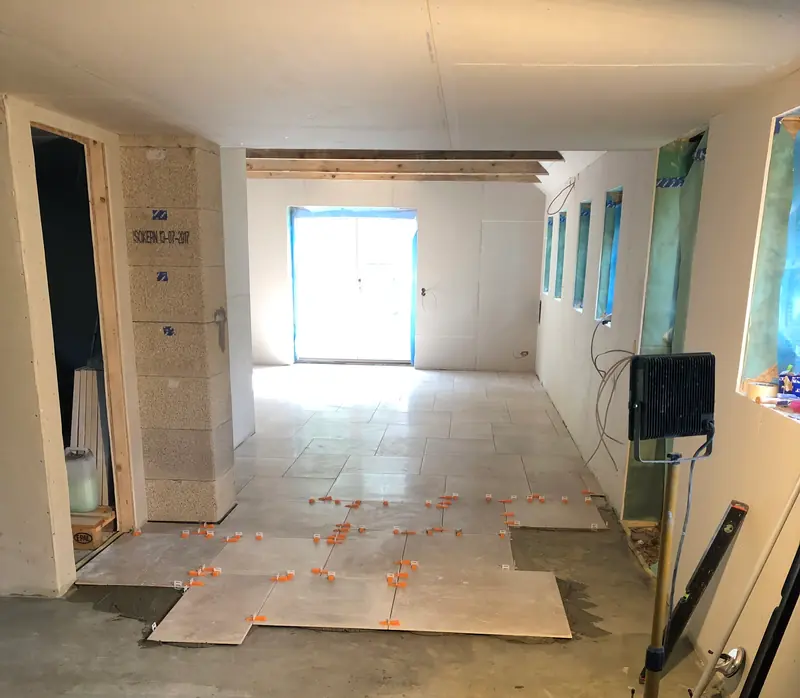 Stort tomt rum under renovering, hvor gulvfliserne lægges ud fra døråbningen med orange fugekryds og pap som afdækning