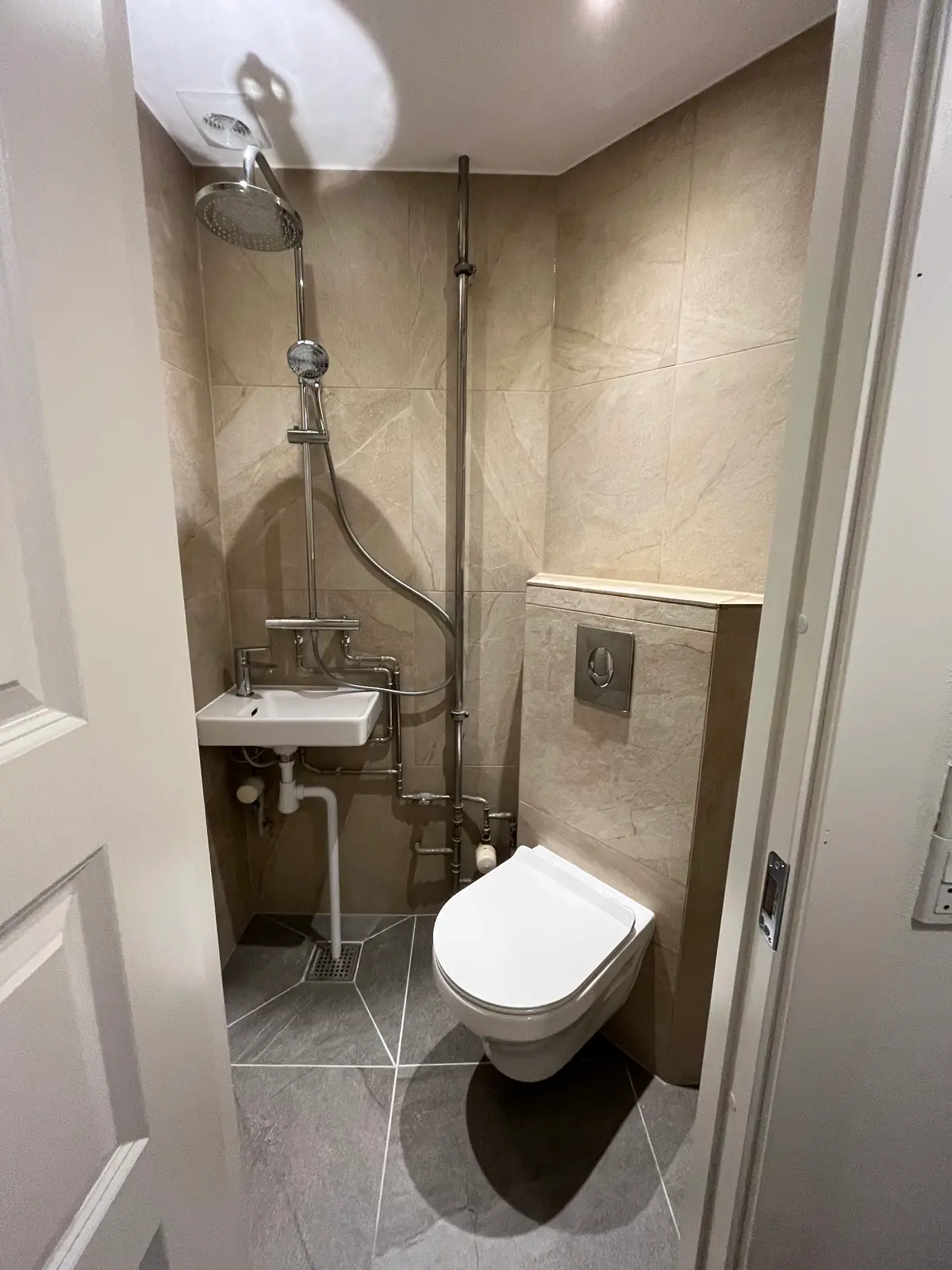 Det færdige minibad med beige vægfliser, grå diagonale gulvfliser, flisehylde over indbygningsvæggen og hvidmalet loft
